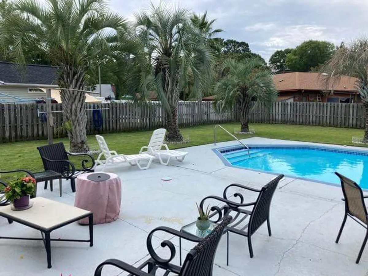 A broad view of the backyard pool area, showing multiple lounge chairs, lush palm trees, and patio furniture. The scene evokes an inviting, leisurely mood, hinting at both daytime sunbathing and evening gatherings under the stars.