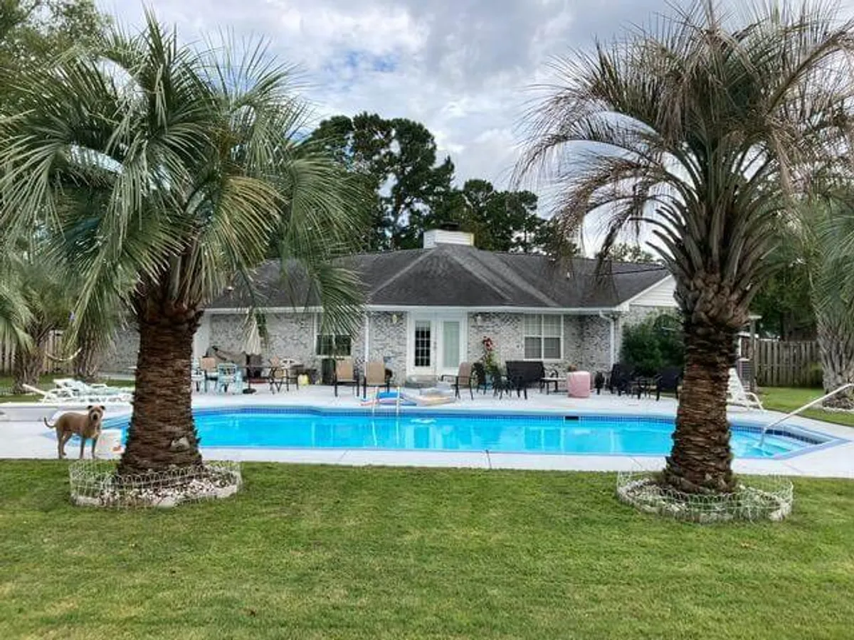 This image features a sparkling inground swimming pool framed by palm trees and patio furniture, capturing a lively yet relaxed outdoor atmosphere perfect for both daytime fun and evening relaxation. The fenced yard ensures privacy, while the pool area is clearly designed for social gatherings and family enjoyment.