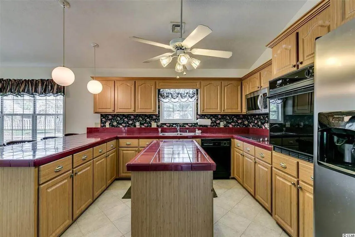 A warm and inviting kitchen scene centers on a striking red countertop island, with patterned curtains and contrasting cabinetry. The arrangement encourages group cooking, entertaining, and shared experiences in a homey environment.