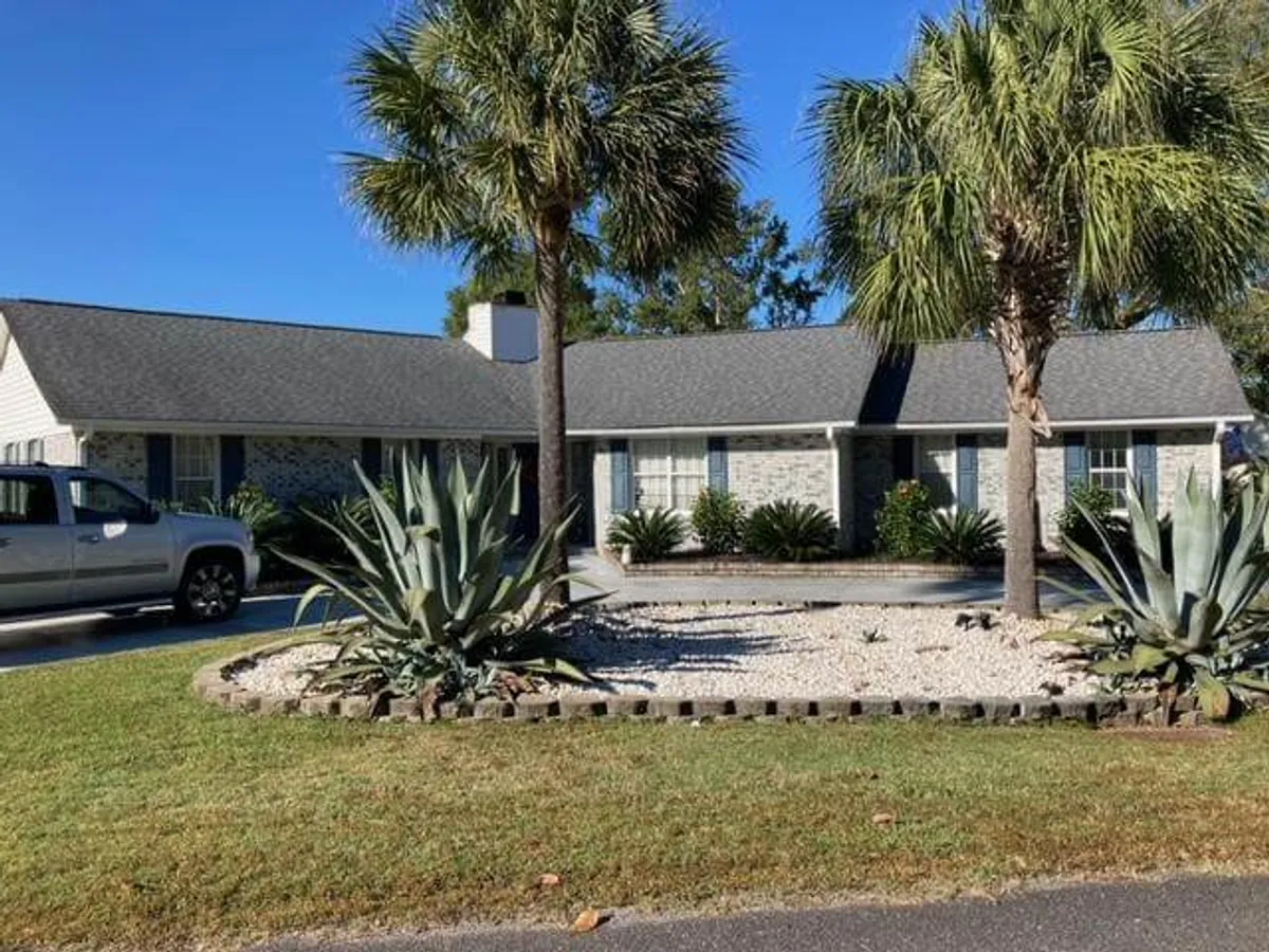 The image showcases the front exterior of 23 Cimmeron Dr, with lush palm trees, succulent plants, a welcoming front porch, attractive stone landscaping, and a vehicle parked in the driveway. The scene conveys a sense of spaciousness and hints at ample parking and storage for vehicles like boats or motorcycles.