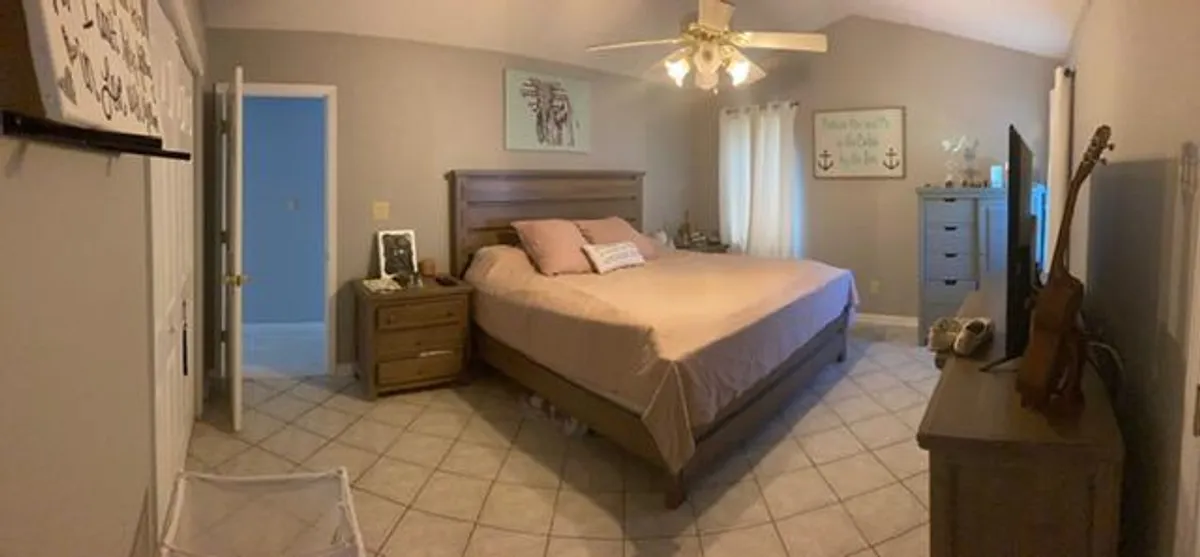 A bright and inviting bedroom with a comfortable bed, nightstand, dresser, wall-mounted television, and a guitar in the corner. Sunlight streams in, creating a peaceful atmosphere ideal for relaxation or family moments.