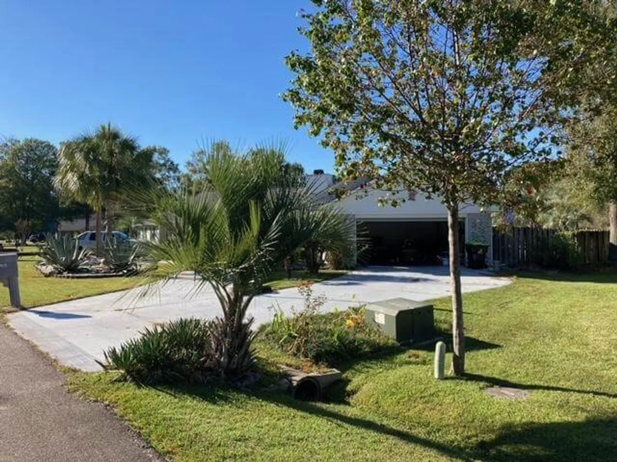 This image presents a clear view of the driveway and attached garage, framed by lush landscaping, palm trees, and a flowering shrub. The scene communicates both accessibility and a sense of welcome, with the driveway in the foreground leading directly to the secure garage.
