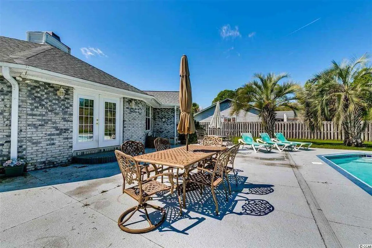 This image showcases a spacious backyard featuring a large swimming pool surrounded by palm trees. An outdoor dining table sits poolside, suggesting space for family barbecues and gatherings. The relaxed atmosphere is enhanced by the golden hour sunlight, perfect for both daytime pool play and evening relaxation.